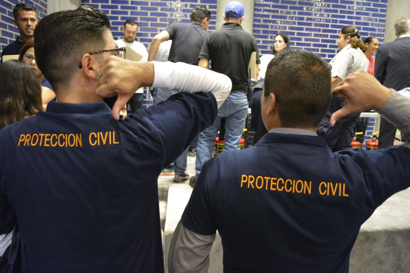 Dos personas señalando a su espalda con su pulgar. En su camisa se lee "PROTECCION CIVIL".